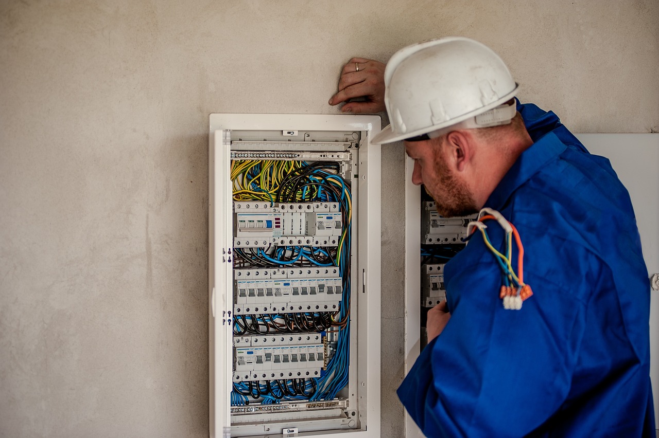 An electrician in a hard hat and coveralls working at an open switchboard.
